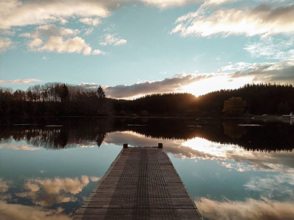 Looking out from a wooden dock to water with trees and setting sun in the distance.